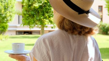 Hasır şapkalı mutlu kadın elinde bir fincan sıcak içecek ve kahve tutuyor, güneşli bir günde yaz terasında oturuyor. Çay içerken görgülü ol. Günaydın, seyahat konsepti.