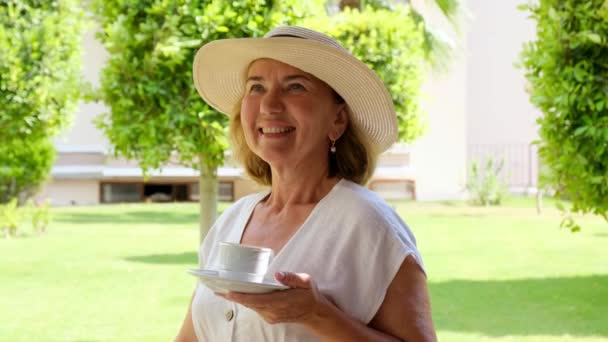 Glimlachende volwassen vrouw van 50-55 jaar in een strohoed drinkt 's morgens vroeg op het terras van haar huis in een groene tuin op een zonnige dag een warme borrel koffie of thee uit een wit kopje. Senior senior — Stockvideo