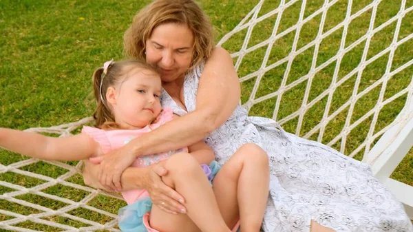 Grandmother and granddaughter, 3 years old, rest and bask in a hammock on a green meadow in their garden on a sunny summer day. Vacation concept, generational relationship, parenting, happy childhood. — Stock Photo, Image