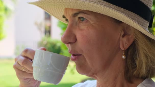 Ενήλικη γυναίκα ηλικίας 50-55 ετών σε ψάθινο καπέλο πίνει ένα ζεστό ρόφημα καφέ ή τσαγιού από ένα λευκό φλιτζάνι νωρίς το πρωί στη βεράντα του σπιτιού της σε έναν καταπράσινο κήπο σε μια ηλιόλουστη μέρα, κοντινό πλάνο. Ανώτερος ανώτερος — Αρχείο Βίντεο