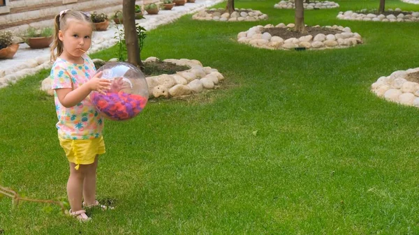 Schattig klein meisje, 3 jaar oud, met twee paardenstaarten op haar hoofd, het dragen van een multi-gekleurde T-shirt en gele shorts, speelt met een multi-gekleurde bal op een groene weide in de tuin van haar huis. De — Stockfoto
