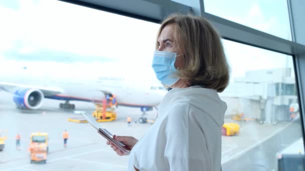 Взрослая женщина в защитной маске стоит у окна терминала аэропорта в ожидании вылета рейса из-за ограничений на перелет в связи с пандемией коронавируса, пожилой человек в возрасте 50-55 лет проводит — стоковое видео