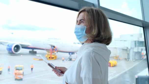 Een volwassen vrouw met een beschermend masker staat bij het raam van de luchthaventerminal in afwachting van het vertrek van een vlucht als gevolg van reisbeperkingen als gevolg van de coronaviruspandemie, een oudere van 50-55 jaar houdt — Stockvideo