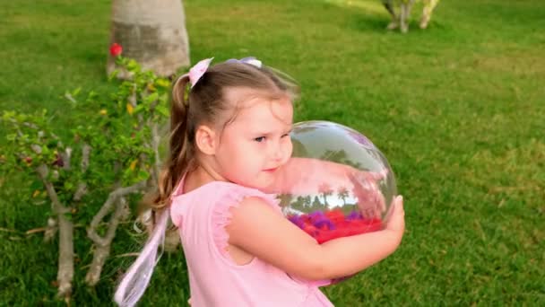 Krásná legrační holčička, 3 roky, se dvěma culíky na hlavě, oblečená v jemných a mnohobarevných šatech růžové modré barvy, hraje s jasnou průhlednou koulí s multi-barevné — Stock video