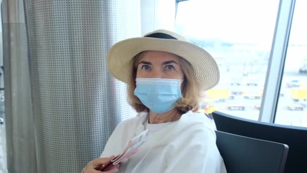 Kobieta w wieku 50-55 lat, w kapeluszu i masce ochronnej, siedzi przy kasie odpraw w terminalu lotniska, czekając na odlot, trzymając bilety lotnicze z powodu podróży — Wideo stockowe