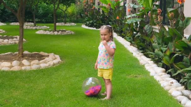 머리에 두 개의 꼬리를 갖고 있는 3 세의 귀여운 소녀는 다양 한 색상의 티셔츠와 노란 반바지를 입고 집 정원에 있는 녹색 초원에서 다양 한 색깔의 공을 가지고 놀고 있다. 미국 의 — 비디오