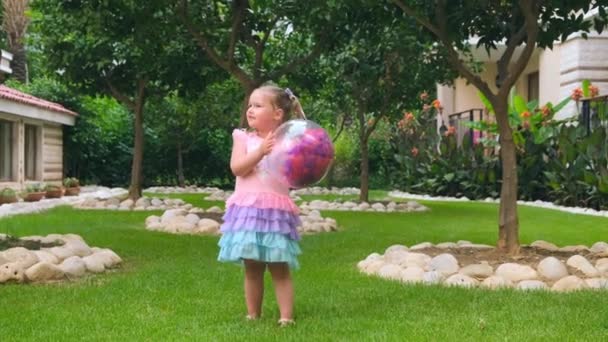 Menina engraçada, 3 anos, com dois rabos-de-cavalo na cabeça, vestida com um vestido delicado e multicolorido de cor azul rosa, joga com uma bola transparente brilhante com penas multicoloridas — Vídeo de Stock