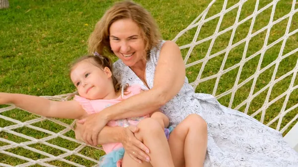 Funny couple grandmother and granddaughter 3 years old are relaxing and basking in a hammock on a green meadow in their garden on a summer sunny day. Vacation concept, generational relationship — Stock Photo, Image