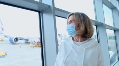 Korunaklı maskeli bir kadın, 50-55 yaş arası, Coronavirus salgını nedeniyle seyahat kısıtlamalarından dolayı havaalanı terminalinin penceresinden uçmayı bekliyor.