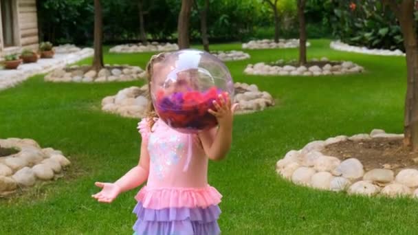 Menina, 3 anos, com dois rabos-de-cavalo na cabeça, vestida com um vestido delicado e multicolorido de cor azul rosa, joga com uma bola transparente brilhante com penas multicoloridas dentro dele — Vídeo de Stock