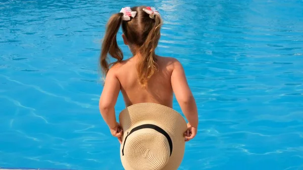 Kleines Mädchen steht und bedeckt sich mit einem Sonnenhut vor dem Hintergrund des blauen Wassers des Pools oder Meeres. Sommerurlaub, Sonnencreme, Reisen. Selektiver Fokus auf Hut — Stockfoto