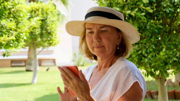 Smutná blondýna senora žena, 50-55 let, v klobouku a bílých šatech, mluví pomocí smartphonu přes videohovor, píše zprávu, hledá informace na internetu na své terase na slunné — Stock fotografie