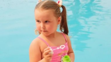 4-5 yaşlarında, pembe mayo giymiş, yaz tatilinde havuzda sabun topu üflüyor.
