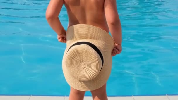 Niña está de pie y se cubre con un sombrero de sol contra el telón de fondo del agua azul de la piscina o el mar. El concepto de vacaciones de verano, protectores solares, viajes. enfoque selectivo en el sombrero — Vídeo de stock