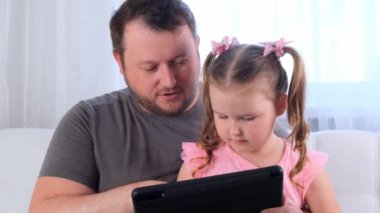 Üç yaşında gülen küçük bir kız ve babası birlikte tablet okuyor ve evde online bir ders izliyorlar. Babam kızına çalışmasında yardım ediyor. Çevrimiçi okul uzaklığı öğrenme kavramı.