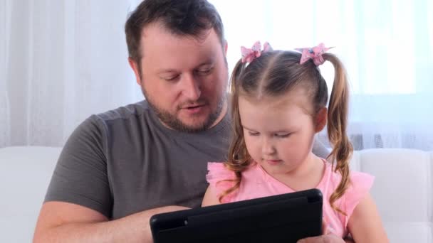 Niña riendo 3 años de edad y su padre estudian juntos en una tableta y ver una lección en línea en casa. Papá ayuda a su hija con sus estudios. Escuela en línea concepto de aprendizaje a distancia. — Vídeo de stock