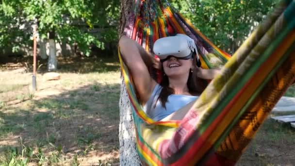 Mulher vestindo óculos de realidade virtual repousa em uma rede perto de uma árvore, imaginando um sonho tornado realidade, um mundo inventado. O conceito de obter experiência usando óculos VR-headset de realidade virtual — Vídeo de Stock
