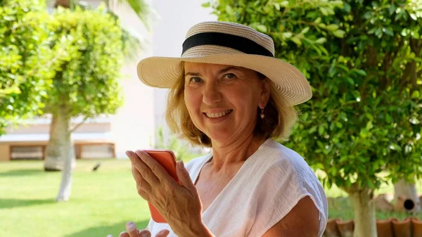 Szőke senora nő, 50-55 éves, kalapban és fehér ruhában, telefonnal beszél videohívással, üzenetet ír, információt keres az interneten a teraszán egy napos reggelen — Stock Fotó