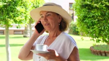 Sarışın bayan, 50-55 yaşlarında, şapkalı ve beyaz elbiseli, akıllı telefon kullanarak konuşuyor ve güneşli bir yaz sabahında terasında elinde bir fincan kahve tutuyor. Serbest konsept