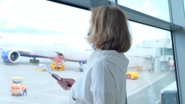 Een volwassen vrouw met een beschermend masker staat bij het raam van de luchthaventerminal in afwachting van het vertrek van een vlucht als gevolg van reisbeperkingen als gevolg van de coronaviruspandemie, een oudere van 50-55 jaar houdt — Stockvideo