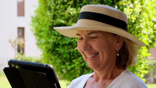 Sorridente anziana donna bionda donna in cappello utilizza tablet pc, tablet digitale per lavoro d'affari o di studio nel suo giardino verde. donna tra i 50 e i 55 anni. Il concetto di propria impresa e formazione — Video Stock