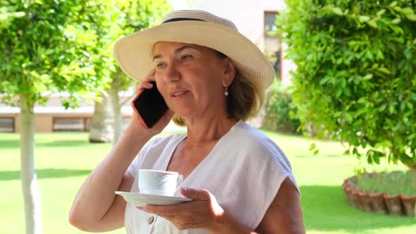 Блондинка-сеньора, 50-55 лет, в шляпе и белом платье, разговаривает с помощью смартфона и держит чашку кофе в руках на террасе солнечным утром в летний день. Концепция фриланса — стоковое видео