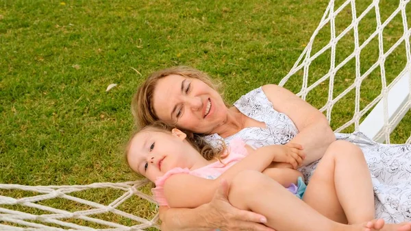 Cheerful Couple grandmother and granddaughter 3 years old are relaxing and basking in a hammock on a green meadow in their garden on a summer sunny day. Vacation concept, generational relationship — Stock Photo, Image