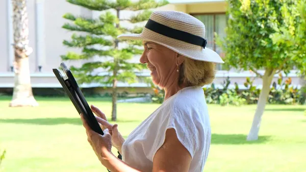 Seniorin blonde Geschäftsfrau mit Hut nutzt Tablet-PC, digitales Tablet für geschäftliche Arbeit oder Studium in ihrem eigenen grünen Garten. Frauen zwischen 50 und 55 Jahren. Das Konzept des eigenen Geschäfts und der Ausbildung, online — Stockfoto