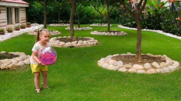 陽気かわいい女の子, 3歳,彼女の頭の上に2つのポニーテールと,マルチカラーのTシャツと黄色のショートパンツを身に着けています,彼女の庭の緑の草原でマルチカラーのボールで遊んでいます — ストック動画
