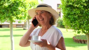 Sarışın bayan, 50-55 yaşlarında, şapkalı ve beyaz elbiseli, akıllı telefon kullanarak konuşuyor ve güneşli bir yaz sabahında terasında elinde bir fincan kahve tutuyor. Serbest konsept