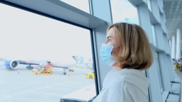Korunaklı maskeli bir kadın, 50-55 yaş arası, Coronavirus salgını nedeniyle seyahat kısıtlamalarından dolayı havaalanı terminalinin penceresinden uçmayı bekliyor. — Stok video
