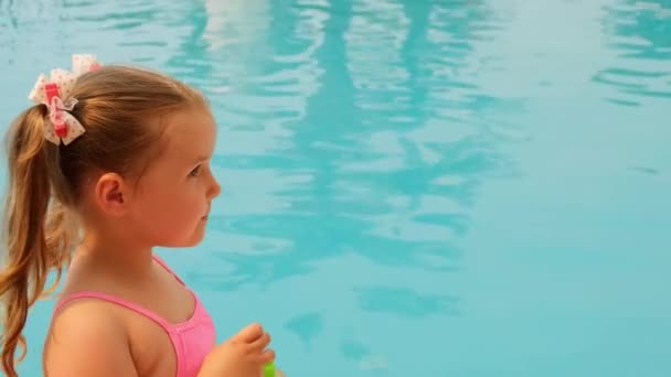 Klein meisje 4-5 jaar oud, gekleed in een roze badpak, blazen zeep ballen in het zwembad op zomervakantie — Stockvideo