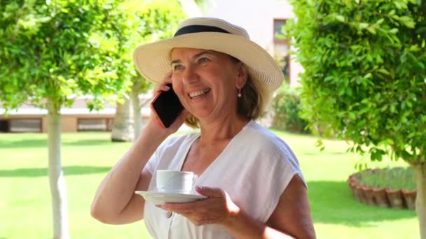 Blondynka seniora, 50-55 lat, w kapeluszu i białej sukience, rozmawia za pomocą smartfona i trzyma filiżankę kawy w dłoniach na tarasie w słoneczny poranek w letni dzień. Koncepcja wolnego strzelca — Wideo stockowe