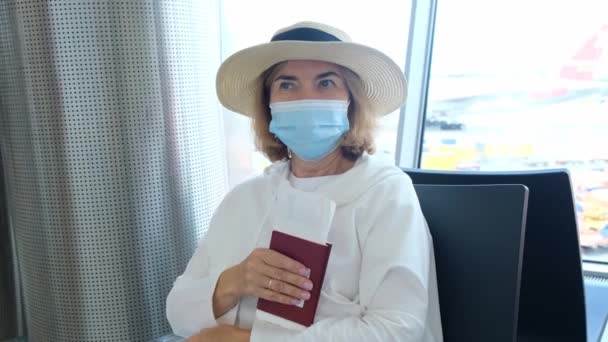 Femmina di 50-55 anni, con cappello e maschera protettiva, si siede ai banchi del check-in del terminal aeroportuale, in attesa della partenza del volo, in possesso di biglietti aerei a causa del viaggio — Video Stock