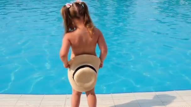 Маленька дівчинка стоїть і вкривається сонячним капелюхом на тлі блакитної води басейну або моря. Концепція літніх канікул, сонцезахисних кремів, подорожей. вибірковий фокус на капелюсі — стокове відео