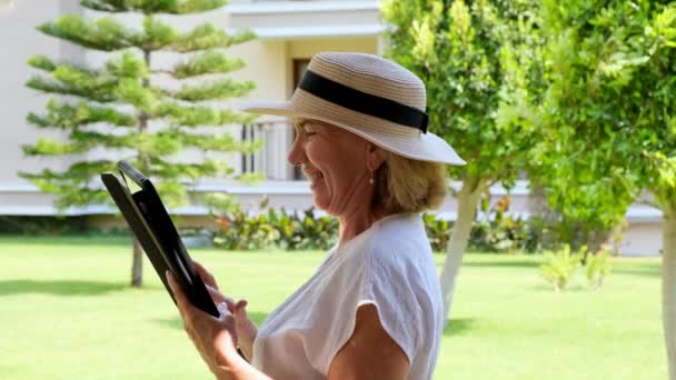 Femme d'affaires blonde senior vif chapeau utilise tablette pc, tablette numérique pour le travail d'affaires ou étudier dans son propre jardin vert. femme âgée de 50 à 55 ans. Le concept de leur propre entreprise et formation — Video