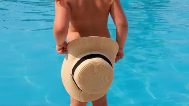 Little girl stands and covers herself with a sun hat against the backdrop of the blue water of the pool or sea. The concept of summer vacations, sunscreens, travel. selective focus on hat — Stock Video