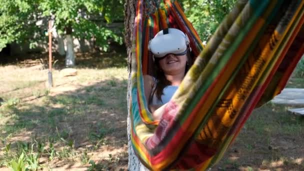 Смешная женщина в очках виртуальной реальности лежит в гамаке рядом с деревом, представляя, как сбывается мечта, выдуманный мир. Концепция получения опыта с помощью виртуальных гарнитур — стоковое видео