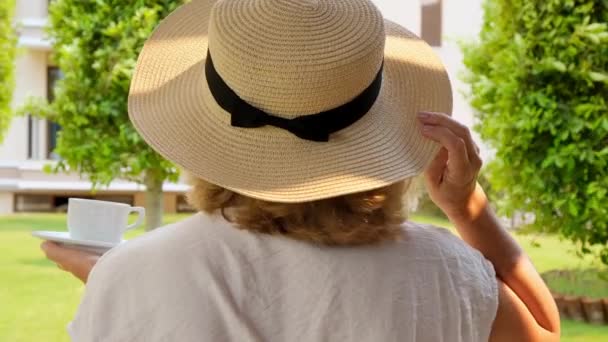 Senora vrouwtje, leeftijd 50-55 in een strohoed houdt een kopje warme drank en drinkt koffie, zittend in de ochtend op een zomerterras op een zonnige dag. Het concept van goedemorgen, reizen en verwachting van — Stockvideo