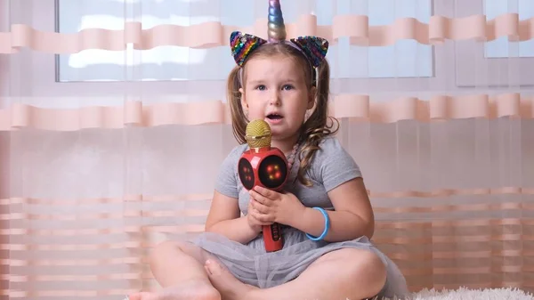 Ελκυστικό τρελό παιδί κυρία τραγουδούν σε ένα μικρόφωνο καραόκε, με ένα μονόκερο headband, το παιδί τραγουδά μουσική καραόκε, διασκεδάστε σε μια εκδήλωση στο μέλλον μουσικός δυνατή φωνή σόλο — Φωτογραφία Αρχείου