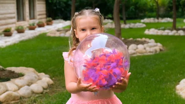 3歳の少女で、頭に2本のポニーテールがあり、ピンクブルーの色の繊細で多色のドレスを着て、その中に複数の色の羽を持つ明るい透明なボールで遊んでいます — ストック写真