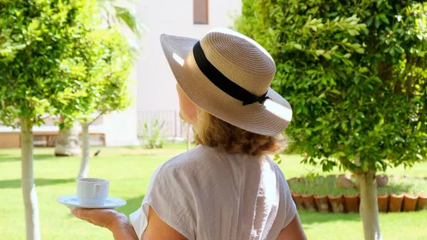 Женщина в возрасте 50-55 лет в соломенной шляпе держит чашку горячего напитка и пьет кофе, сидя утром на летней террасе в солнечный день. Концепция доброго утра, путешествия и ожидания нового — стоковое фото