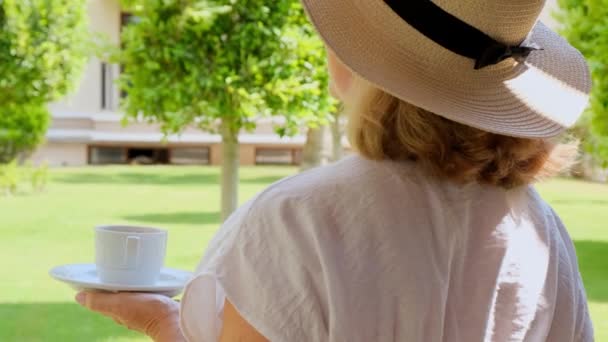 Kobieta w wieku 50-55 lat w słomkowym kapeluszu trzyma filiżankę gorącego napoju i pije kawę, siedząc rano na letnim tarasie w słoneczny dzień. Pojęcie dzień dobry, podróże i oczekiwania nowych — Wideo stockowe