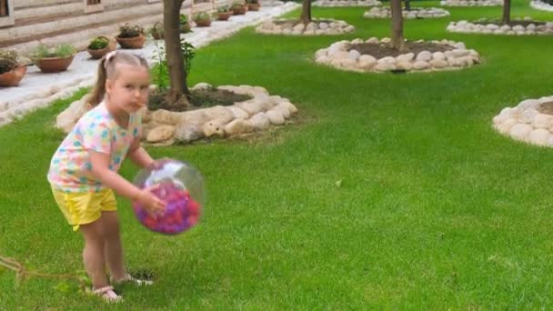 Смішна дівчинка, 3 роки, з двома хвостами на голові, одягнена в різнокольорову футболку і жовті шорти, грає з різнокольоровим м'ячем на зеленому лузі в саду — стокове відео