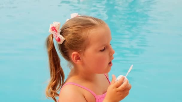 Joyeuse petite fille de 4-5 ans, avec des queues de cheval, portant un maillot de bain rose, soufflant des boules de savon dans la piscine en vacances d'été — Video