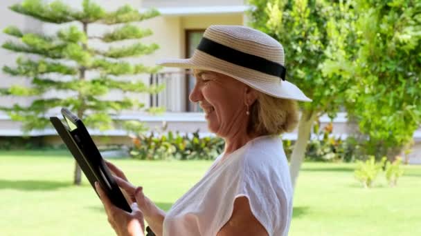 Starsza blondynka bizneswoman w kapeluszu używa tabletu, tabletu cyfrowego do pracy w biznesie lub nauki w swoim zielonym ogrodzie. kobieta w wieku 50-55 lat. Koncepcja własnego biznesu i szkolenia, online — Wideo stockowe