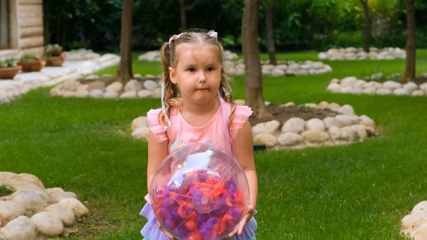 Menina engraçada, 3 anos, com dois rabos-de-cavalo na cabeça, vestida com um vestido delicado e multicolorido de cor azul rosa, joga com uma bola transparente brilhante com penas multicoloridas — Fotografia de Stock