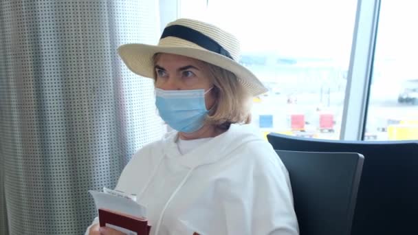 Een volwassen vrouw met een hoed en een beschermmasker zit aan de incheckbalie in de terminal van de luchthaven in afwachting van het vertrek van de vlucht met vliegtickets als gevolg van reisbeperkingen als gevolg van de — Stockvideo