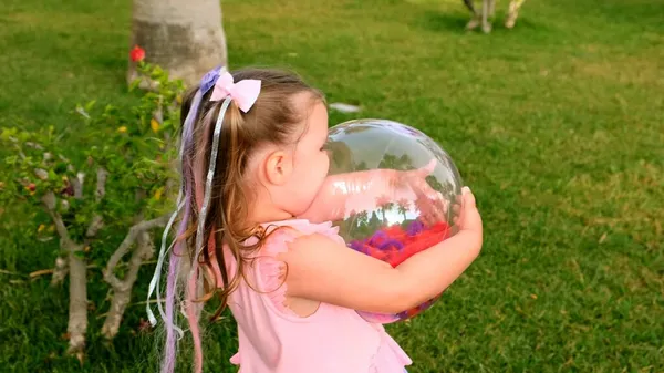 Mooi grappig klein meisje, 3 jaar oud, met twee paardenstaarten op haar hoofd, gekleed in een delicate en multi-gekleurde jurk van roze blauwe kleur, speelt met een heldere transparante bal met multi-gekleurde — Stockfoto
