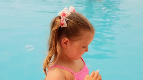 Joyeuse petite fille de 4-5 ans, avec des queues de cheval, portant un maillot de bain rose, soufflant des boules de savon dans la piscine en vacances d'été — Video